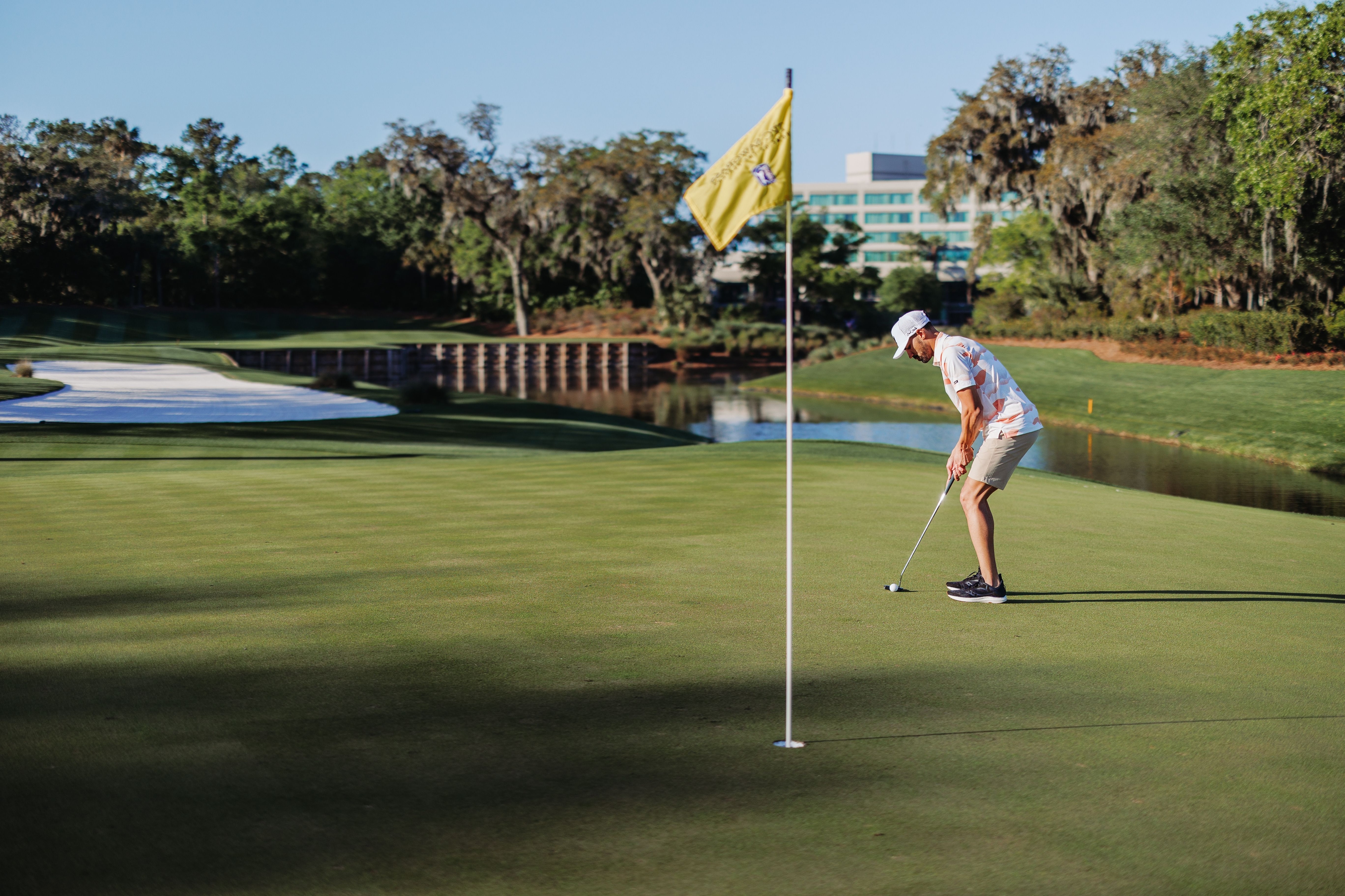 Golfer in a flowery shirt putting on a lush sawgrass green under clear skies, focusing on the ball near the hole on a well-manicured course