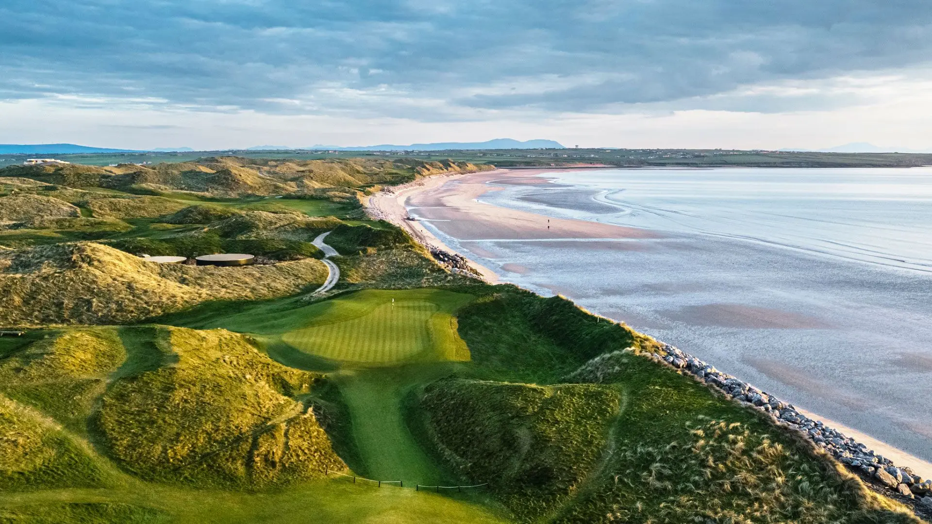 Scenic view of Ballybunion Golf Course with rolling green fairways, dunes, and the Atlantic Ocean coastline under a partly cloudy sky