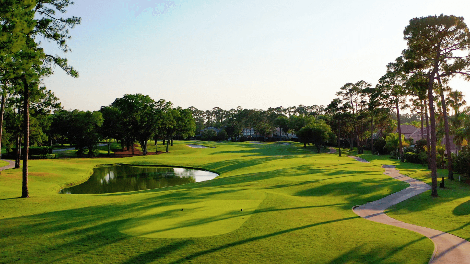 Wide view of a lush green plantation fairway lined with trees under a clear sky, showing the golf hole stretching into the distance